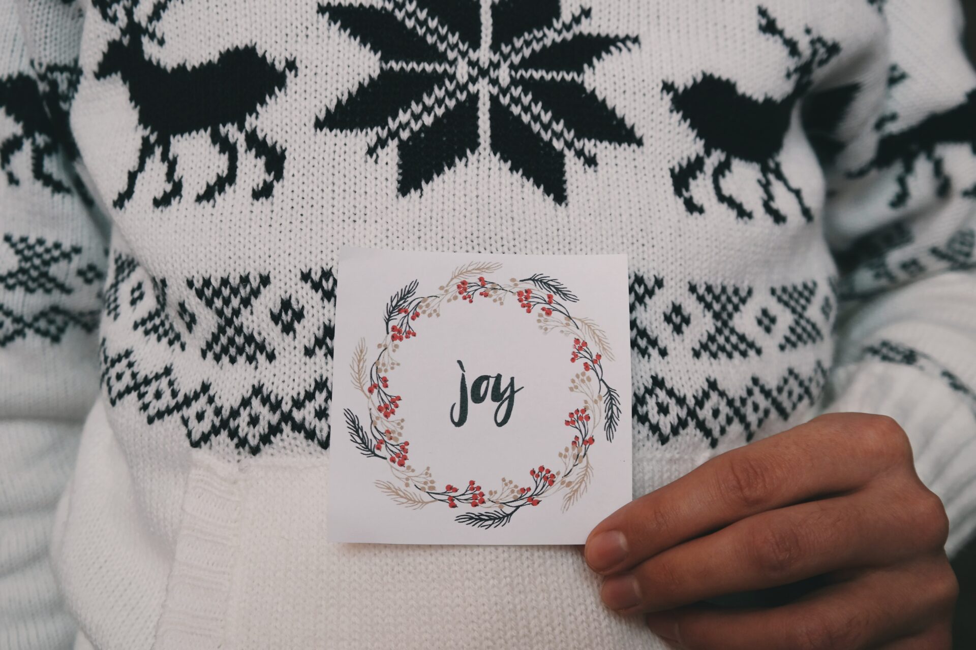 Close up of a man in a white Christmas jumper holding a Christmas card that has the word Joy on it.